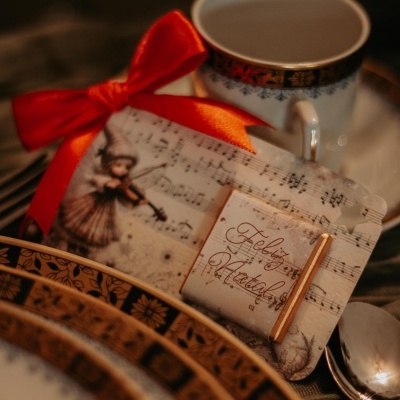 Presente decorativo com notas musicais e anjo, laÃ§o vermelho, etiqueta Feliz Natal, junto a porcelana branca com detalhes dourados