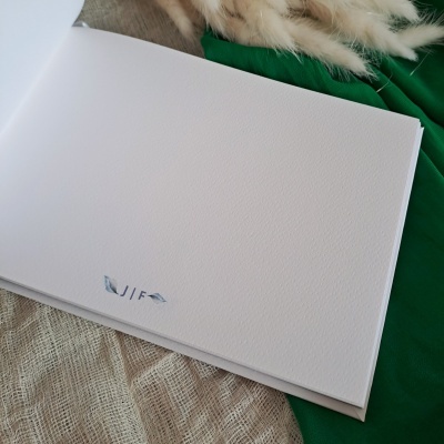 Livro ou álbum branco aberto com letras 'JIF' em relevo sobre tecido bege e verde com plumas brancas.