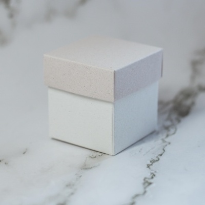 Caixa pequena quadrada com tampa bege e corpo branco sobre mármore branco.