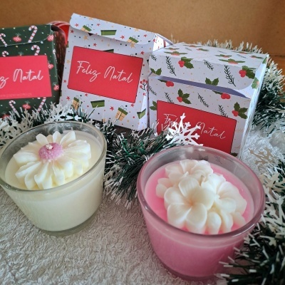 Duas velas decorativas em copos de vidro com flores no topo e caixas natalícias com a frase 'Feliz Natal'