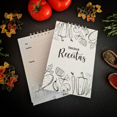 Livro de receitas com capa branca decorada por desenhos a preto de vegetais, rodeado por ingredientes culinários