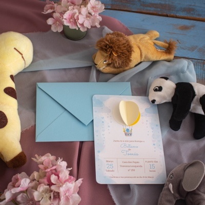 Convite de batizado com envelope azul claro entre peluches e flores sobre tecido colorido