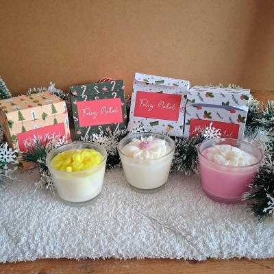 Três velas em copos de vidro com flores de cera no topo e caixas de Natal ao fundo com etiqueta 'Feliz Natal'.