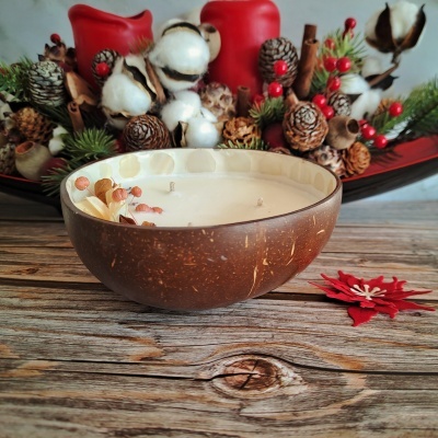 Vela aromática em casca de coco com decoração natural sobre mesa de madeira com arranjo natalício ao fundo.