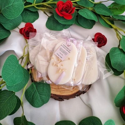 Sabonetes ovais brancos decorados com flores secas e fitas lilás em embalagem transparente sobre suporte de madeira e fundo com folhas e flores artificiais