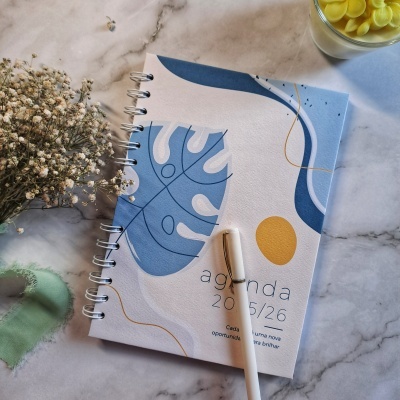 Agenda com capa azul, branca e amarela, caneta branca, flores secas e copo com pastilhas amarelas