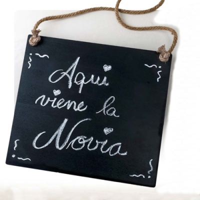 Placa preta com texto 'Aquí viene la Novia' em giz branco pendurada por corda