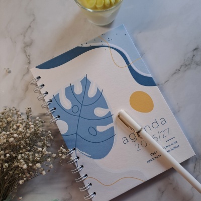 Caderno espiral com padrão azul e branco, caneta e planta seca sobre mesa de mármore