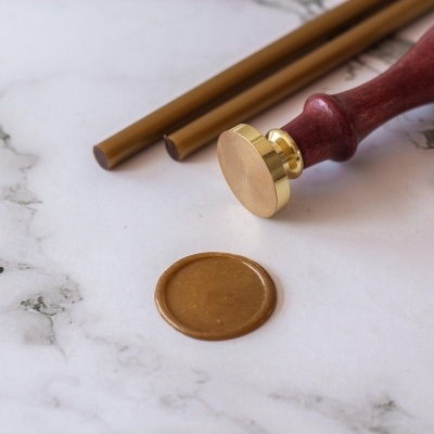 Selo de lacre dourado com cabo de madeira e varetas douradas sobre superfície marmoreada