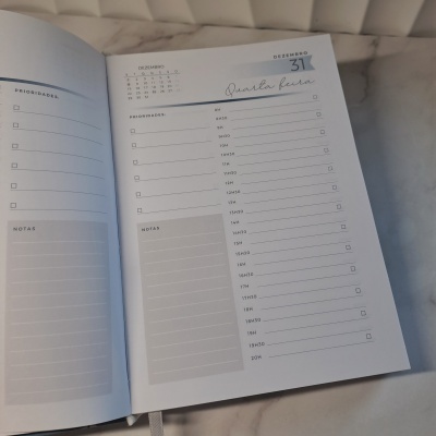 Página aberta de planner diário com datas e horários, detalhes em azul e branco, fundo de mármore