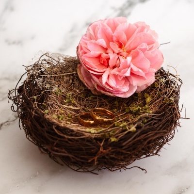 Ninho decorativo com flor rosa e anéis dourados sobre superfície marmorizada branca