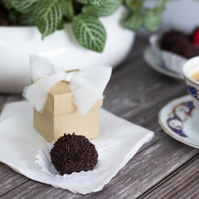 Brigadeiro e caixa de presente com laço branco sobre mesa de madeira com xícara de café e planta ao fundo
