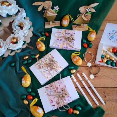 Caixas brancas decoradas, coelhos de madeira, ovos decorativos, canetas brancas e pulseiras em exibidos sobre tecido verde e mesa de madeira.