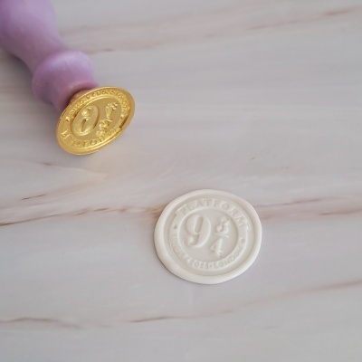 Carimbo de lacre com cabo roxo e selo dourado ao lado de selo branco com texto em relevo