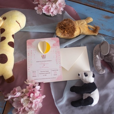 Convite de nascimento rosa com balão amarelo e branco, rodeado por peluches de girafa, leão, elefante e panda sobre mesa azul