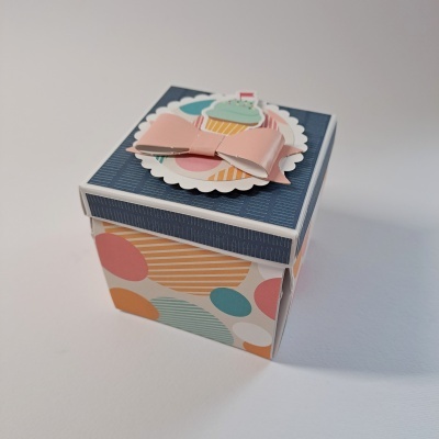 Caixa quadrada decorada com padrão colorido e tampa azul com enfeite de cupcake e laço rosa