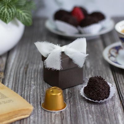 Caixa de presente castanha com laço branco, bombom com granulado, cápsula de café dourada e livro sobre mesa de madeira.