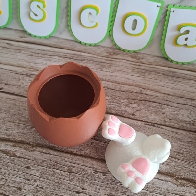 Caixa em forma de ovo castanho com tampa de coelho branca e patas cor-de-rosa, sobre mesa de madeira e faixa decorativa verde e branca ao fundo.