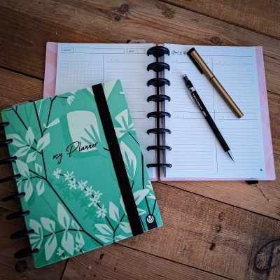 Dois planners em superfície de madeira, um verde com padrão de folhas e outro aberto com caneta e lápis