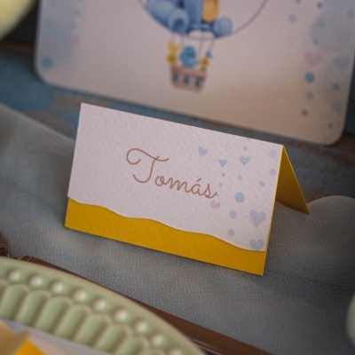 Cartão de nome branco com base amarela e nome 'Tomás' em fonte castanha sobre tecido cinzento