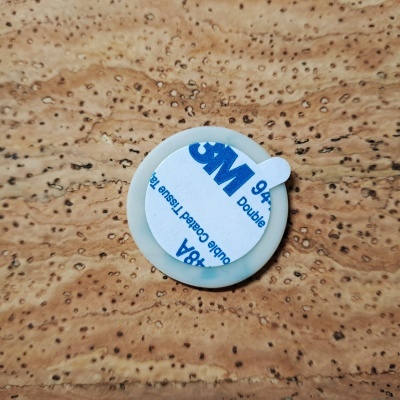 Peça circular branca com adesivo 3M sobre superfície castanha texturizada