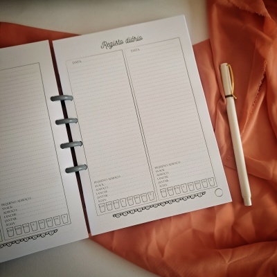 Folhas de registo diário de papel branco com texto cinza, sobre tecido laranja, com caneta branca ao lado