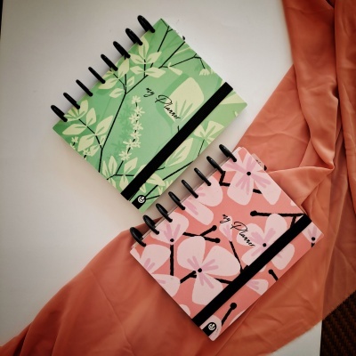 Dois cadernos espiralados com elástico preto e padrões florais, um verde e outro rosa, sobre tecido laranja.