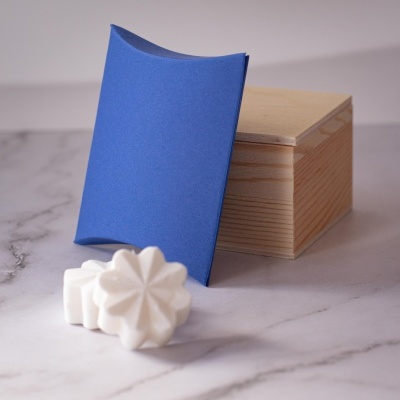 Caixa de madeira clara, embalagem azul curva e pastilhas brancas florais sobre superfície marmoreada.