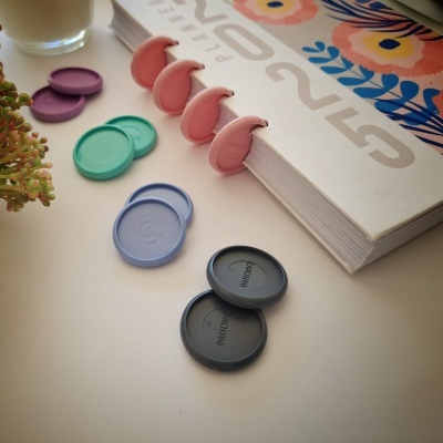 Marcadores de páginas coloridos em plástico sobre mesa branca com livro e planta