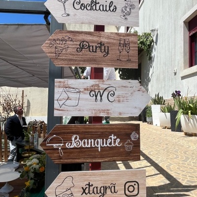 Placas de madeira com setas e texto indicando várias direções para diferentes locais e atividades numa festa ao ar livre.