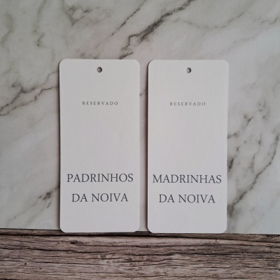 Dois cartões brancos com textos RESERVADO, PADRINHOS DA NOIVA e MADRINHAS DA NOIVA sobre mármore cinza e madeira.