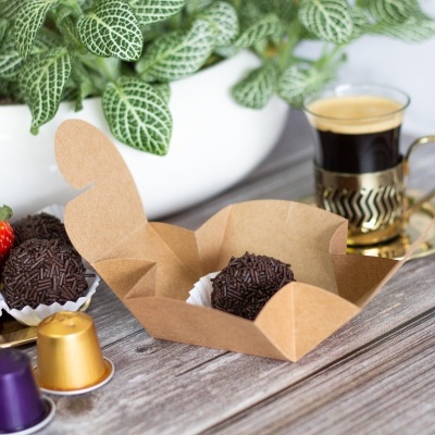 Brigadeiros em caixa de papel castanho, cápsulas de café, chávena de café expresso e planta verde sobre mesa de madeira.