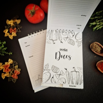 Blocos de receitas com capa branca e ilustrações a preto, rodeados de ingredientes de cozinha