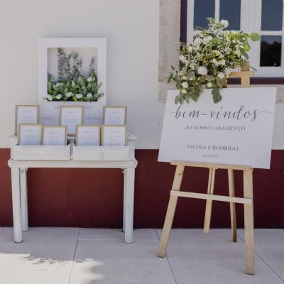 Mesa branca com molduras douradas e cartaz de boas-vindas decorado com flores