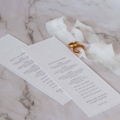 Dois menus brancos com texto em português, fita branca e dois anéis dourados sobre superfície de mármore
