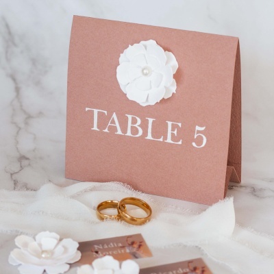 Cartão de mesa castanho com flor branca e texto TABLE 5, com dois anéis dourados e tecido branco