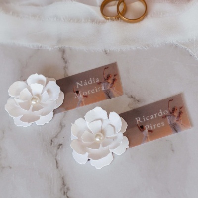 Etiquetas transparentes com nomes e flores brancas em papel com centro em pérola, sobre mármore, com anéis dourados ao fundo