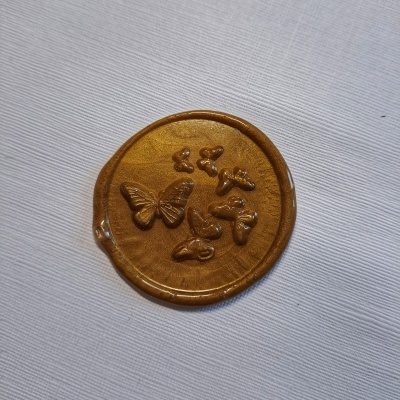 Selo de cera dourada com borboleta e formas pequenas em relevo sobre tecido branco