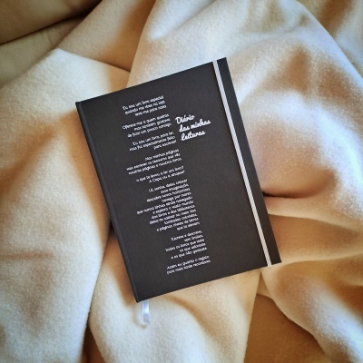 Caderno preto com texto impresso em português, elástico branco e fita marcador, sobre tecido bege acolchoado.