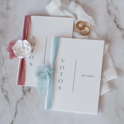 Dois cadernos de votos de casamento com bordas rosa e azul, um com flor branca, outro com fita azul, com o nome RICARDO no caderno azul