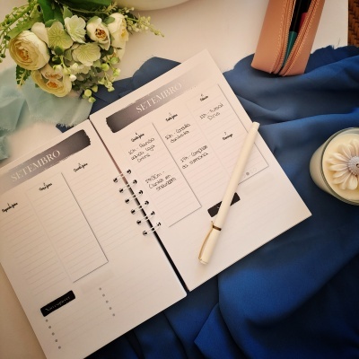 Agenda aberta com caneta branca, flores, estojo rosa e vela decorativa sobre tecido azul