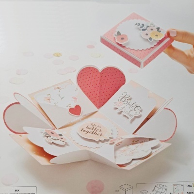 Caixa de presente explodida com decoração floral e corações, texto em inglês, ao lado de uma mão segurando a tampa.