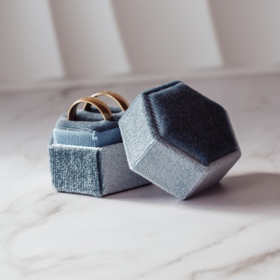 Estojo de veludo cinzento hexagonal com anéis dourados aberto sobre superfície branca