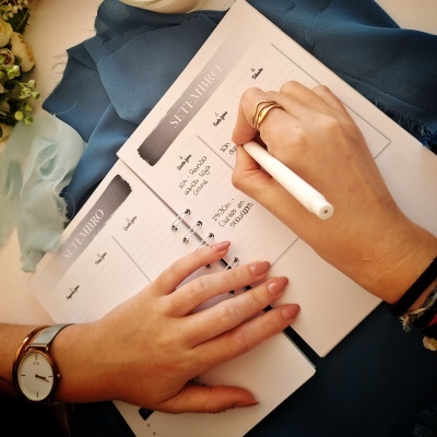 Caderno semanal com caneta e mãos femininas escrevendo, tecido azul e flores ao lado