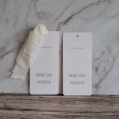 Duas etiquetas em papel branco texturizado com texto RESERVADO MÃE DA NOIVA e RESERVADO MÃE DO NOIVO sobre superfície de mármore e madeira.