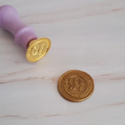 Selo de cera dourado com punho roxo e texto 'PLATFORM 9 3/4 KING'S CROSS LONDON' sobre madeira clara
