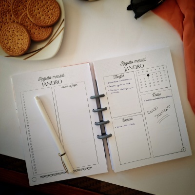 Caderno aberto com plano mensal escrito e caneta branca, ao lado prato com bolachas redondas