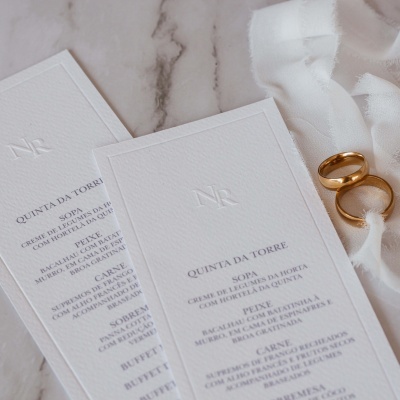 Menus brancos em relevo com texto e dois anéis dourados sobre tecido branco em mármore claro