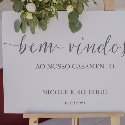 Placa branca retangular com texto de boas-vindas para casamento, decorada com flores e apoiada em cavalete de madeira
