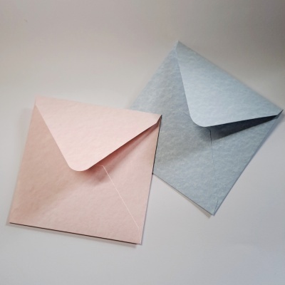Dois envelopes quadrados, um rosa claro e outro azul claro, sobre superfície branca.